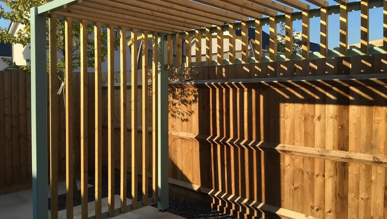 Nowa pergola z drewnianymi listwami i metalową konstrukcją, z cieniami na drewnianym płocie.