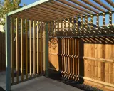 Nowa pergola z drewnianymi listwami i metalową konstrukcją, z cieniami na drewnianym płocie.