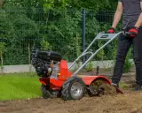 Osoba w rękawiczkach obsługuje czerwoną glebogryzarkę spalinową, która spulchnia ziemię w ogrodzie.