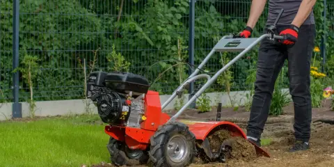 Osoba w rękawiczkach obsługuje czerwoną glebogryzarkę spalinową, która spulchnia ziemię w ogrodzie.