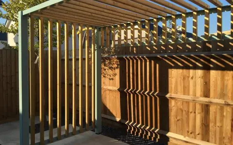 Nowa pergola z drewnianymi listwami i metalową konstrukcją, z cieniami na drewnianym płocie.