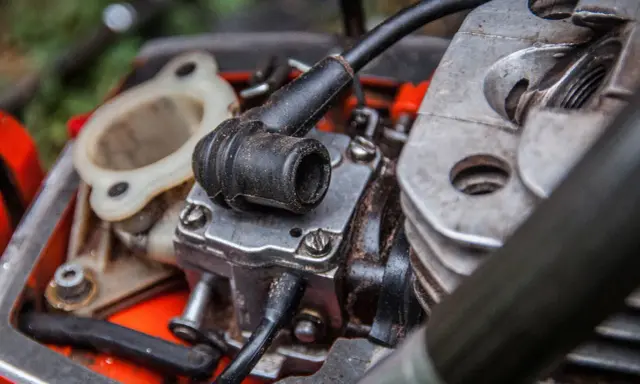 Zbliżenie na silnik kosiarki spalinowej: świeca zapłonowa, gaźnik i cylinder.
