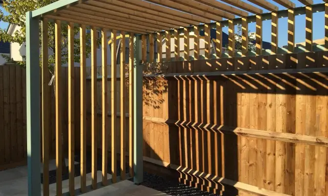 Nowa pergola z drewnianymi listwami i metalową konstrukcją, z cieniami na drewnianym płocie.