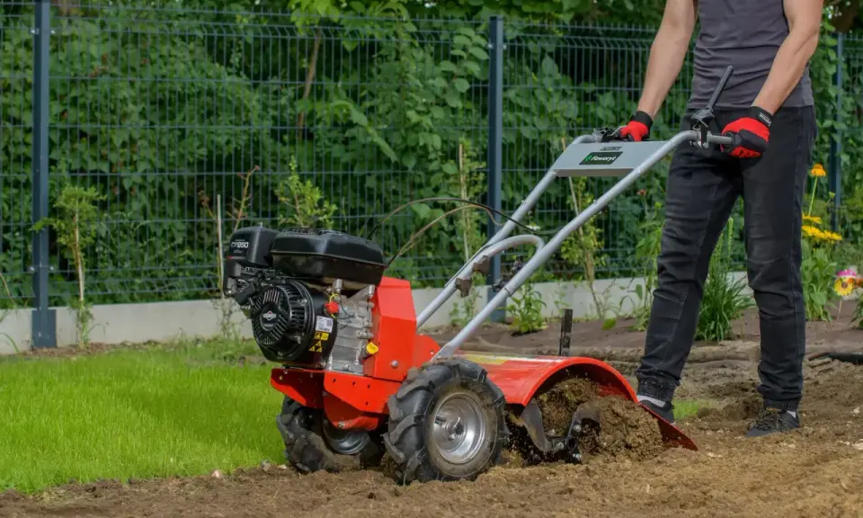 Osoba w rękawiczkach obsługuje czerwoną glebogryzarkę spalinową, która spulchnia ziemię w ogrodzie.